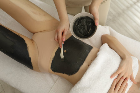 Esthetician applying cosmetic product for body wraps treatment onto woman's belly in spa salon, above viewの写真素材
