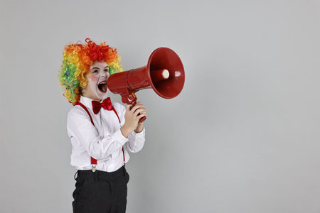Little boy dressed like clown shouting in megaphone on grey background, space for text. Surprise...
