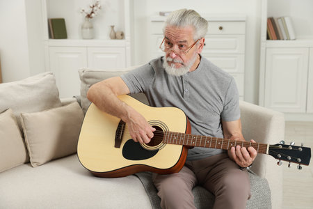 Relaxing hobby. Senior man playing guitar on sofa at homeの写真素材