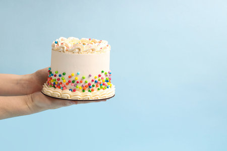 Woman holding delicious cake with colorful sprinkles on light blue background, closeup. Space for textの写真素材