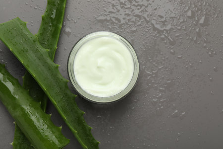 Moisturizing cream with extract of aloe vera in jar, cut leaves and water drops on grey background, flat lay. Space for textの写真素材