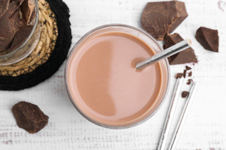 Tasty chocolate milk in glass and pieces of chocolate on white wooden table, flat lay