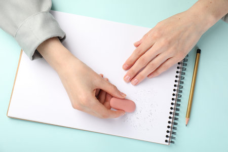 Woman rubbing eraser against paper at light blue table, top view