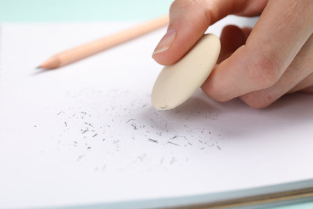 Woman rubbing eraser against paper at light blue table, closeup