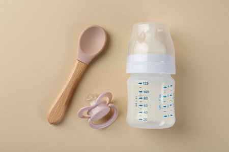 Feeding bottle with milk, spoon and pacifier on beige background, flat lay