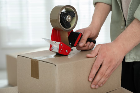Man sealing cardboard box with tape dispenser indoors, closeupの写真素材