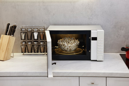 Open microwave oven with bowl of popcorn on white marble countertop in kitchenの写真素材