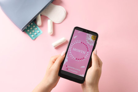 Menstruation. Woman holding smartphone with period calendar application on pink background, top viewの写真素材