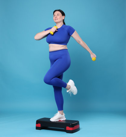Plus size woman in gym clothes doing exercise with dumbbells and step platform on light blue backgroundの写真素材