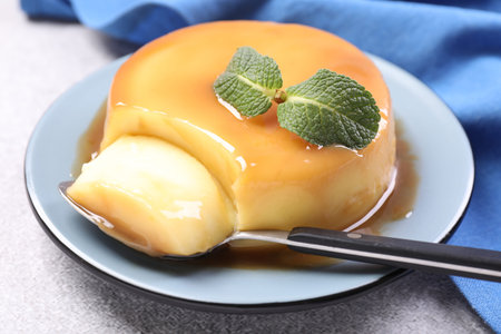 Delicious pudding with caramel sauce, mint and spoon on grey textured table, closeup