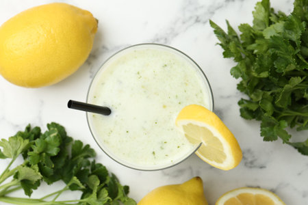 Healthy parsley drink in glass, leaves and lemons on white marble table, flat lay