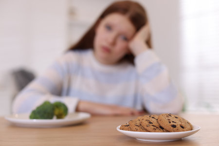 Sad teenage girl with broccoli and cookies at wooden table, selective focus. Eating disorderの写真素材