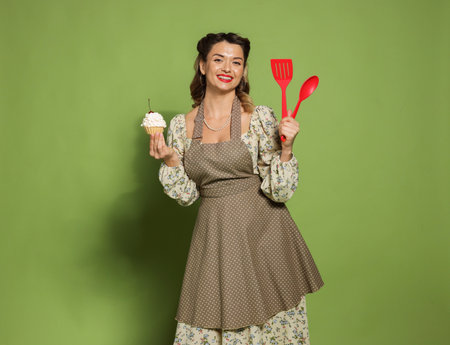 Happy woman with cupcake and cooking utensils on green background. Pin-up vibe portraitの写真素材