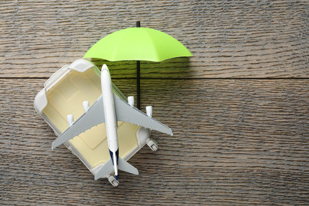 Travel insurance. Plane model, small umbrella and suitcase on wooden table, flat lay. Space for textの写真素材