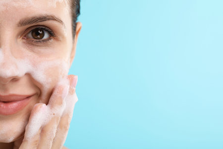 Attractive woman washing her face with cleansing foam on light blue background, closeup. Space for...
