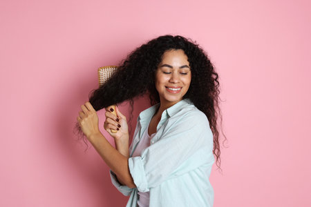Smiling young woman brushing her curly hair on pink backgroundの写真素材