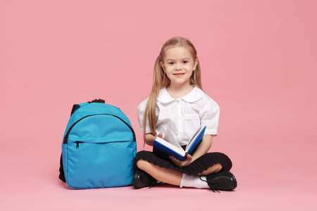 Schoolgirl with backpack reading book on pink background