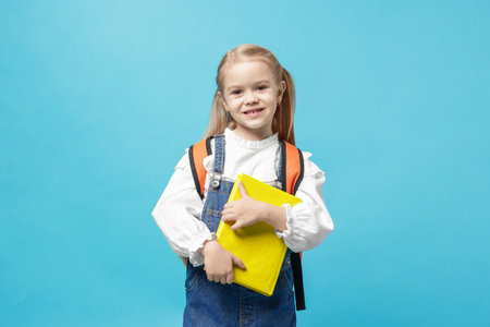 Cute schoolgirl with backpack and book on light blue backgroundの写真素材