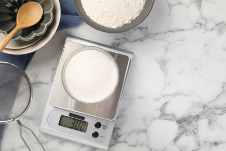 Digital kitchen scale with glass of sugar and other products on white marble table, flat lay. Space for textの写真素材