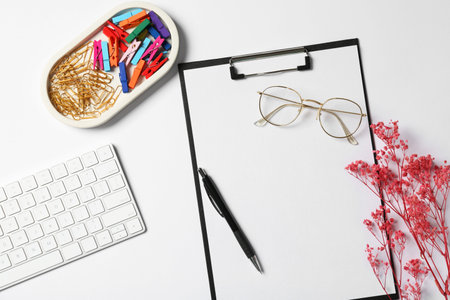 Clipboard, other stationery, flowers, glasses and computer keyboard on white background, flat lay