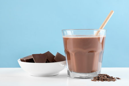 Tasty chocolate milk with shavings and pieces on white table against light blue background
