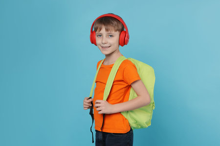 Cute schoolboy in headphones with backpack on light blue background