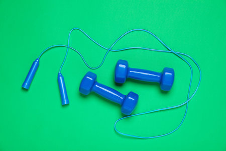 Blue skipping rope and dumbbells on green background, flat lay