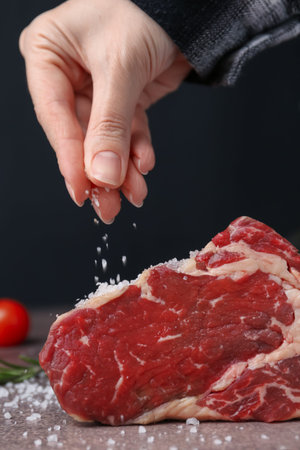 Woman salting piece of raw beef at brown textured table against black background, closeup
