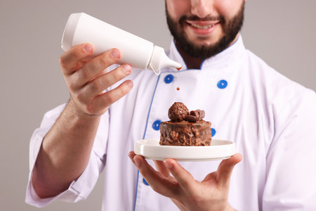 Happy confectioner in uniform holding delicious chocolate dessert and bottle of sauce on light grey background, closeupの写真素材
