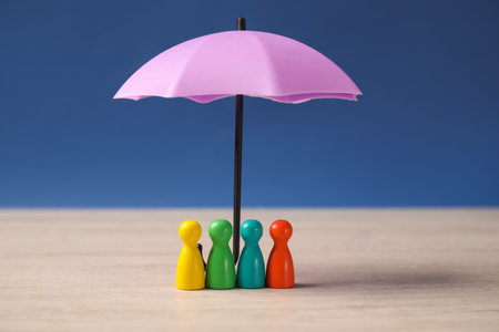 Human figures under umbrella on light wooden table against blue background. Insurance conceptの写真素材