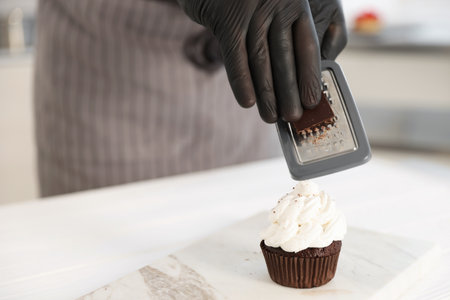 Confectioner grating chocolate onto cupcake at table in kitchen, closeupの写真素材