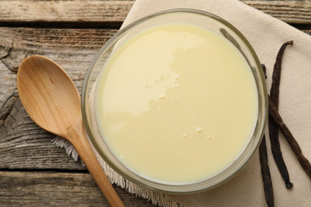 Tasty condensed milk, spoon and vanilla pods on wooden table, flat lay