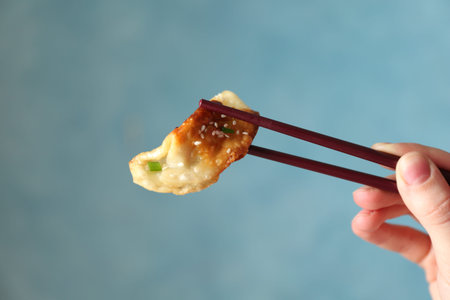 Woman holding tasty fried gyoza (dumpling) with chopsticks on light blue background, closeup