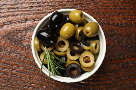 Different delicious marinated olive rings, whole ones and rosemary in bowl on wooden table, top viewの写真素材