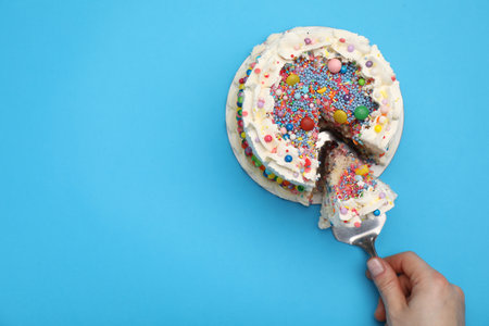 Woman taking piece of tasty cake with colorful sprinkles on light blue background, top view. Space for textの写真素材