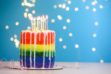 Delicious rainbow birthday cake with burning candles, sprinkles and streamer on light table against blue background with blurred lights, closeup. Space for textの写真素材