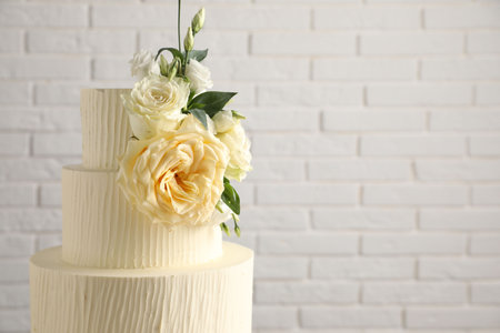 Delicious wedding cake decorated with flowers near white brick wall, closeup. Space for text