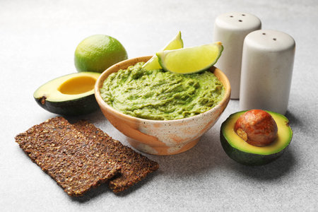Tasty avocado dip, lime and crispbreads on light grey table, closeup
