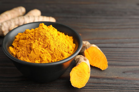 Aromatic turmeric powder in bowl and fresh roots on wooden table, closeup. Space for text