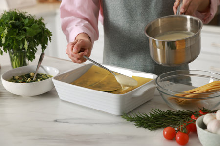 Woman spreading bechamel sauce onto spinach lasagna at marble table indoors, closeupの写真素材