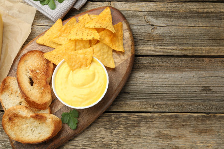 Tasty cheese dipping sauce in bowl, nacho chips and croutons on wooden table, top view. Space for...