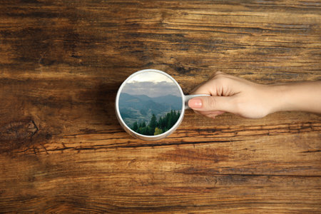 Woman holding ceramic cup with mountain landscape instead of drink at wooden table, top viewの写真素材