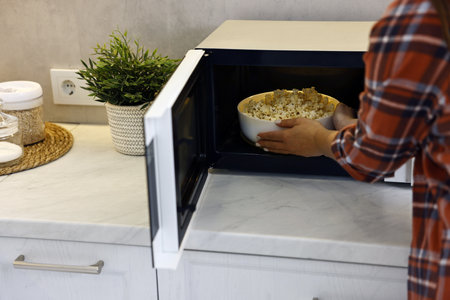 Woman taking bowl with popcorn out of microwave oven at white marble countertop in kitchen, closeupの写真素材