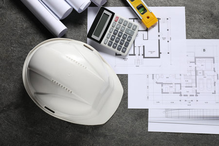Different architectural drawings, hard hat, building level and calculator on gray table, flat lay