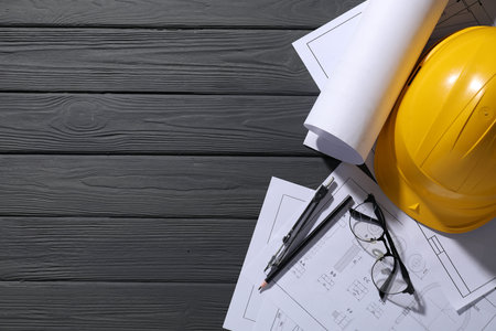 Different engineering drawings, hard hat, glasses, pencil and divider on gray wooden table, flat...