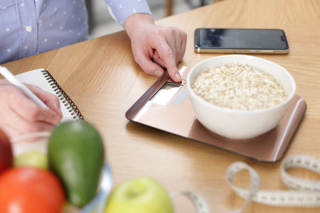 Woman weighing oatmeal and writing in notebook at wooden table, closeupの写真素材