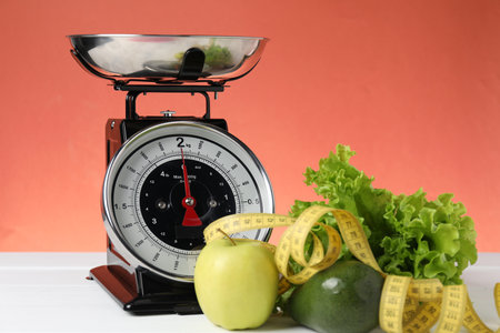 Mechanical kitchen scale with products and measuring tape on white wooden table against coral background, closeup. Dietの写真素材