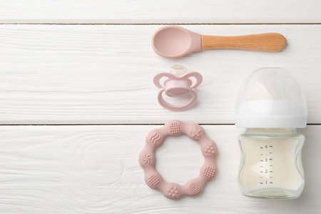 Feeding bottle with milk, spoon, pacifier and teether on white wooden table, flat lay. Space for...