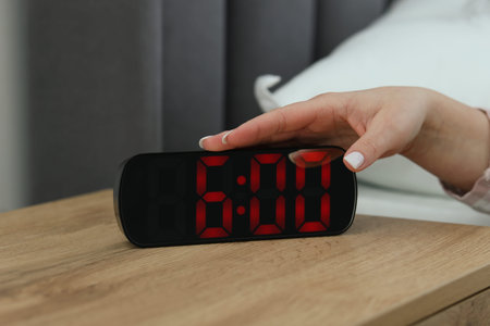 Woman turning off digital alarm clock on bedside table indoors, closeup