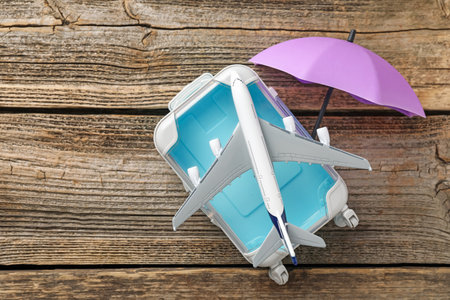 Travel insurance. Plane model, small umbrella and suitcase on wooden table, flat lay. Space for textの写真素材